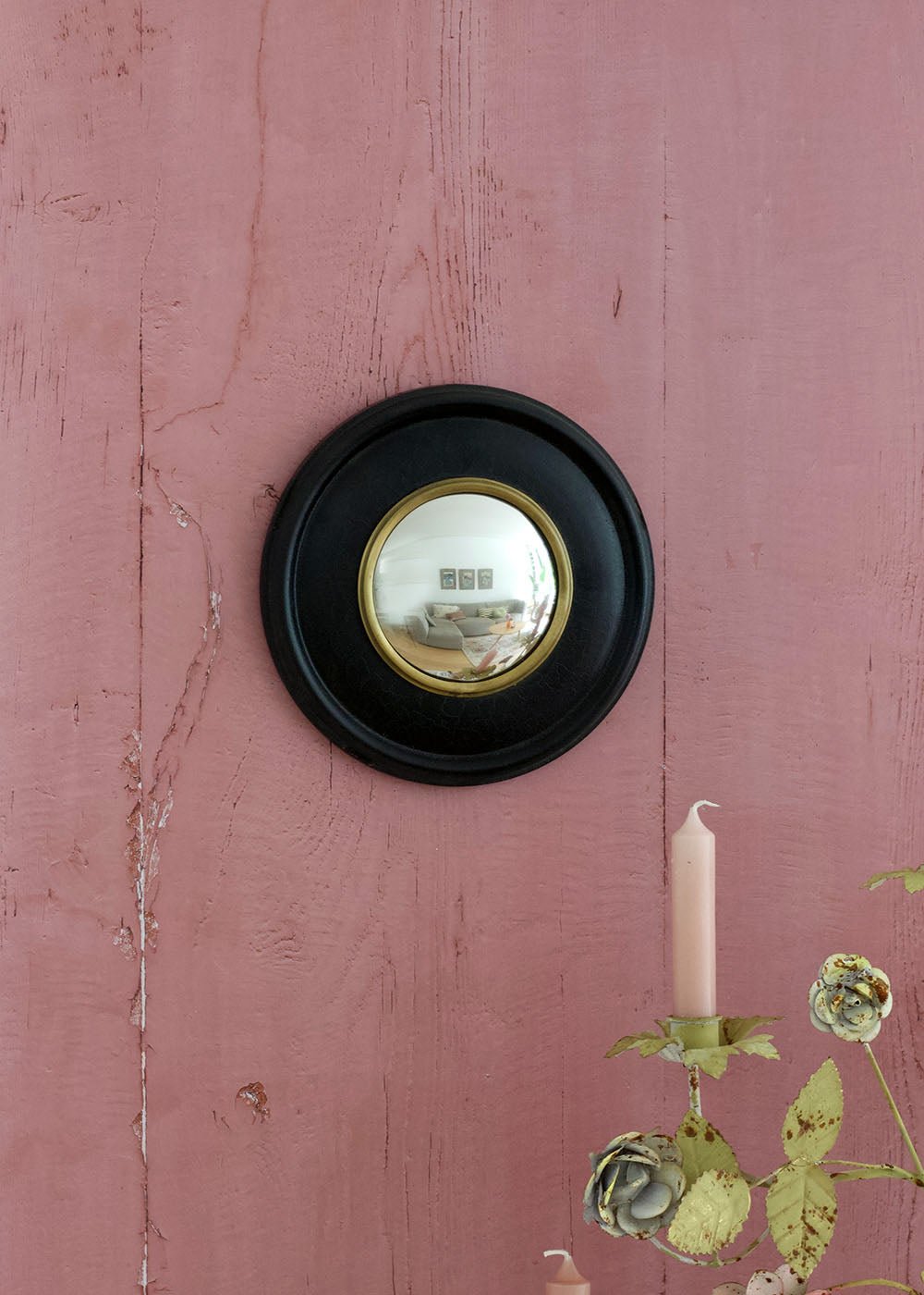 Decorative convex mirror with black frame on a pink wall with a candle and greenery.