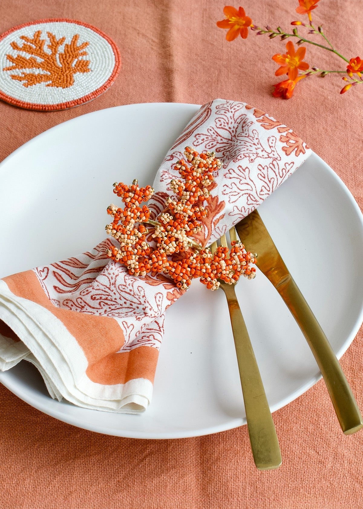 Decorative table setting with orange and white patterned napkin, gold cutlery, and orange berries on a peach-colored tablecloth.