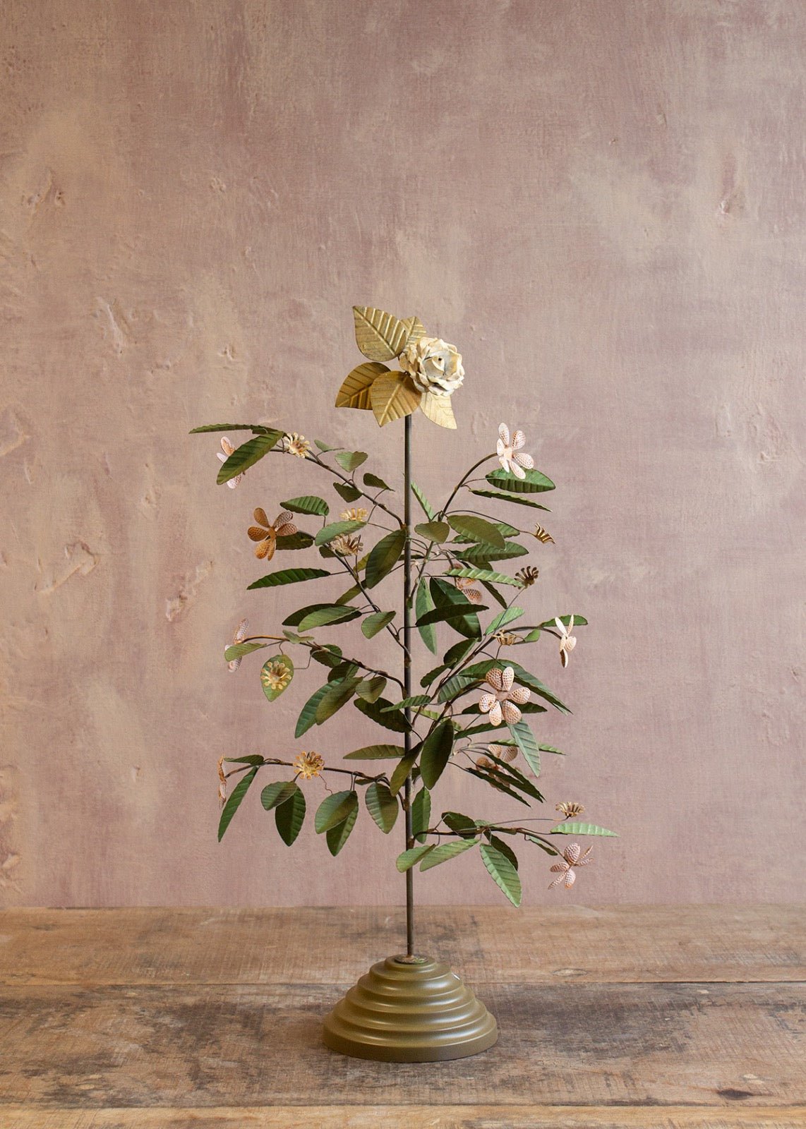 Decorative metal flower tree with green leaves and small flowers on a brass-toned base against a muted pink wall.