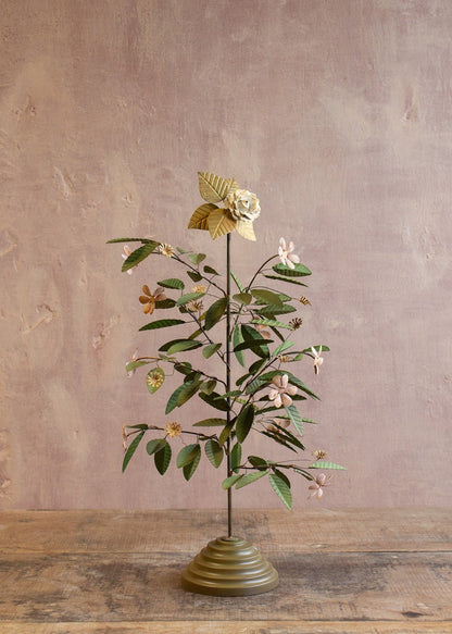 Decorative metal flower tree with green leaves and small flowers on a brass-toned base against a muted pink wall.