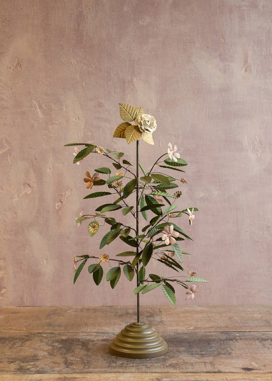 Decorative metal flower tree with green leaves and small flowers on a brass-toned base against a muted pink wall.