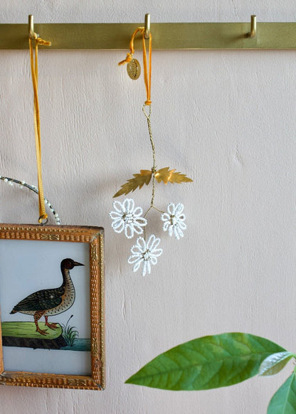 Beaded flower ornament with brass leaves and ochre ribbon hanging on brass hook.
