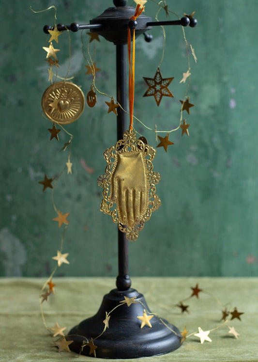 Brass ex-voto hand ornament with decorative frame, hanging on a stand with golden star garland.