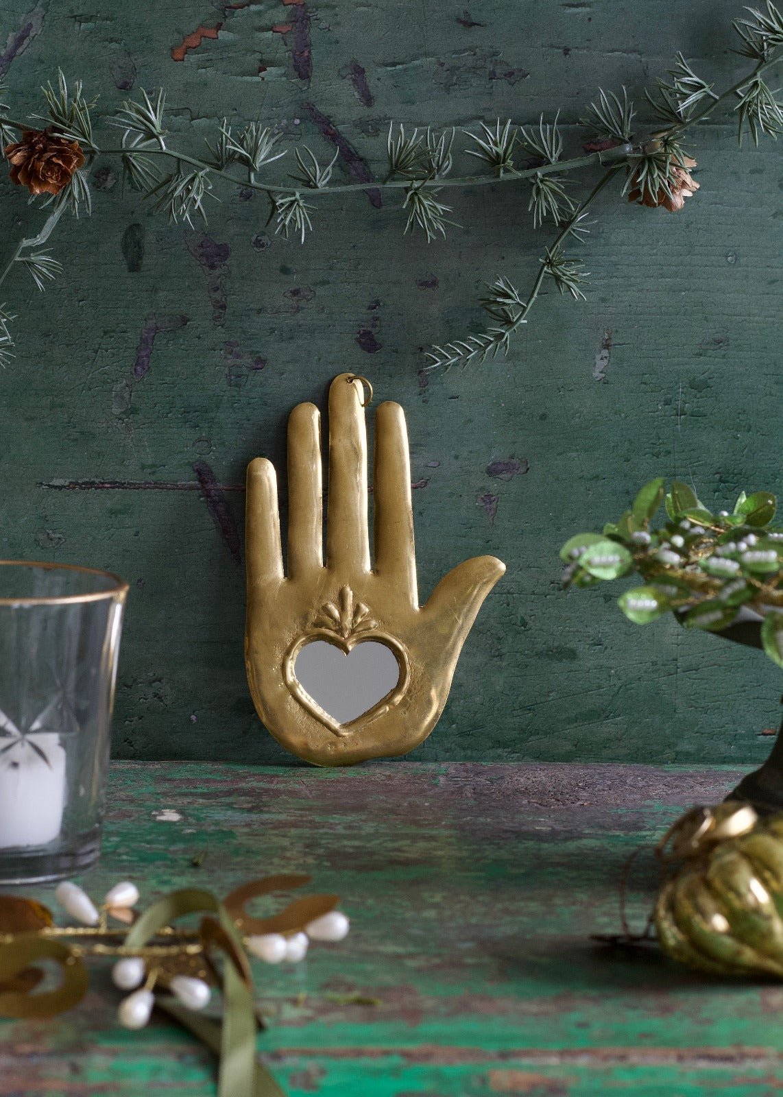 Brass ex-voto hand ornament with a heart-shaped mirror in the palm, displayed against a green background.