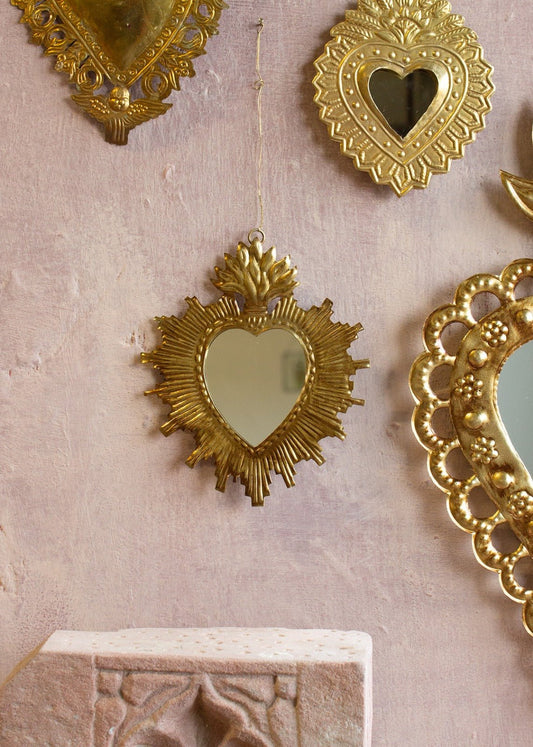 Ex-voto heart mirror with golden radiant frame and flame detail hanging on a pink wall.