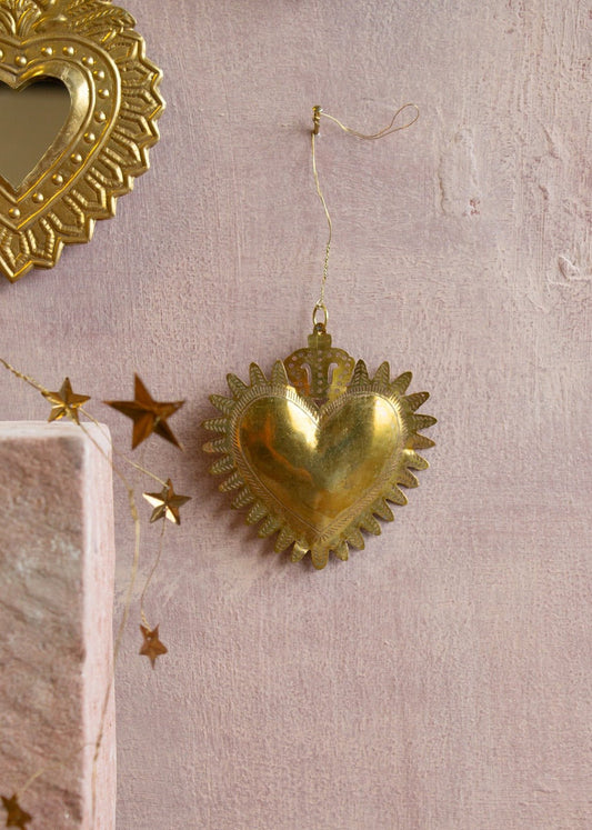 Golden handmade ex-voto heart with embossed crown detail, hanging on a wall with star garland nearby.