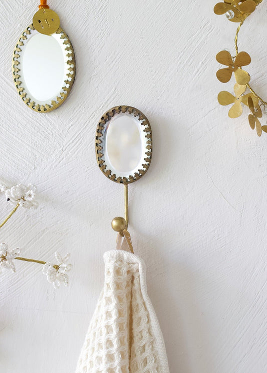 Handmade oval ex voto mirror hook with brass frame, styled on a wall with hanging towel and decorative brass elements.