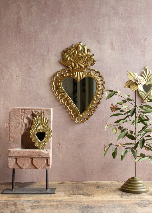 Heart-shaped brass mirror with flame-shaped crown and floral dotted frame, hanging on a pink textured wall with decorative objects and a brass floral tree.