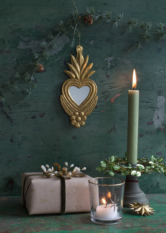 Large heart-shaped ex-voto mirror with golden metal frame, styled on a dark green vintage wall with a lit candle, wrapped gift, and festive green wreath.