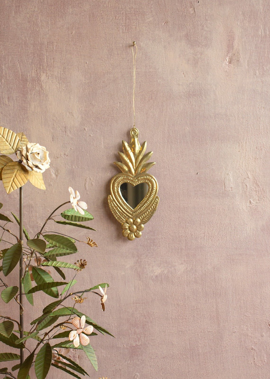 Small gold ex-voto mirror in heart shape, hanging on a soft mauve wall next to a vintage floral metal sculpture.