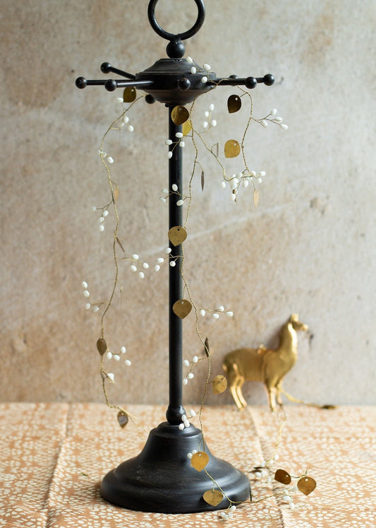 Black metal jewelry stand with a decorative garland of gold heart-shaped leaves and white beads, placed on a patterned cloth with a golden deer figurine in the background
