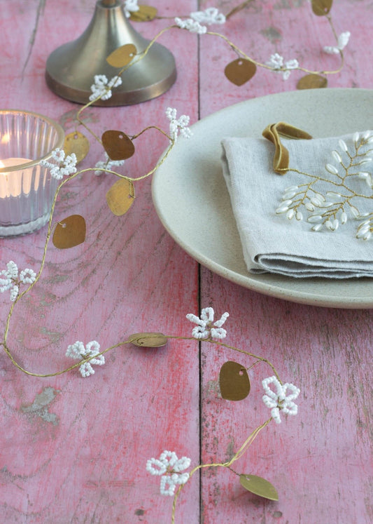 Garland with small white beaded flowers and brass heart-shaped leaves, styled on a rustic pink wooden table next to a candle, linen napkin with gold embroidery, and a ceramic plate.