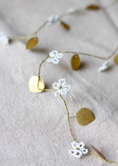 Close-up of a garland featuring white beaded flower blossoms and small gold brass leaves, arranged on a soft beige linen fabric.