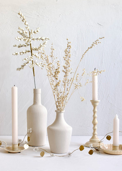 Delicate garland with white beaded flowers and brass leaves arranged across a white surface, styled with cream-toned vases, dried flowers, and tall candles in matching candleholders against a textured white wall.