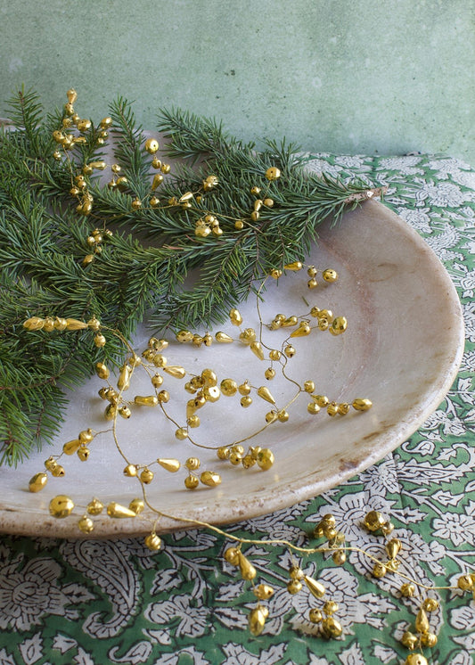 Golden brass garland with faceted teardrop and round beads, styled with fir branches in a marble bowl on a patterned green cloth.