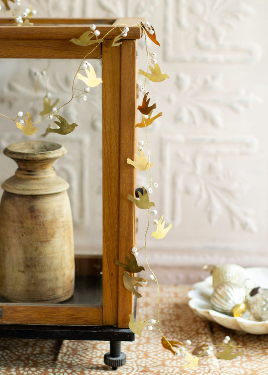 Garland with brass bird silhouettes and small white berries draped along the edge of a wooden glass cabinet, with neutral decor elements and ornaments in the background on a patterned cloth.