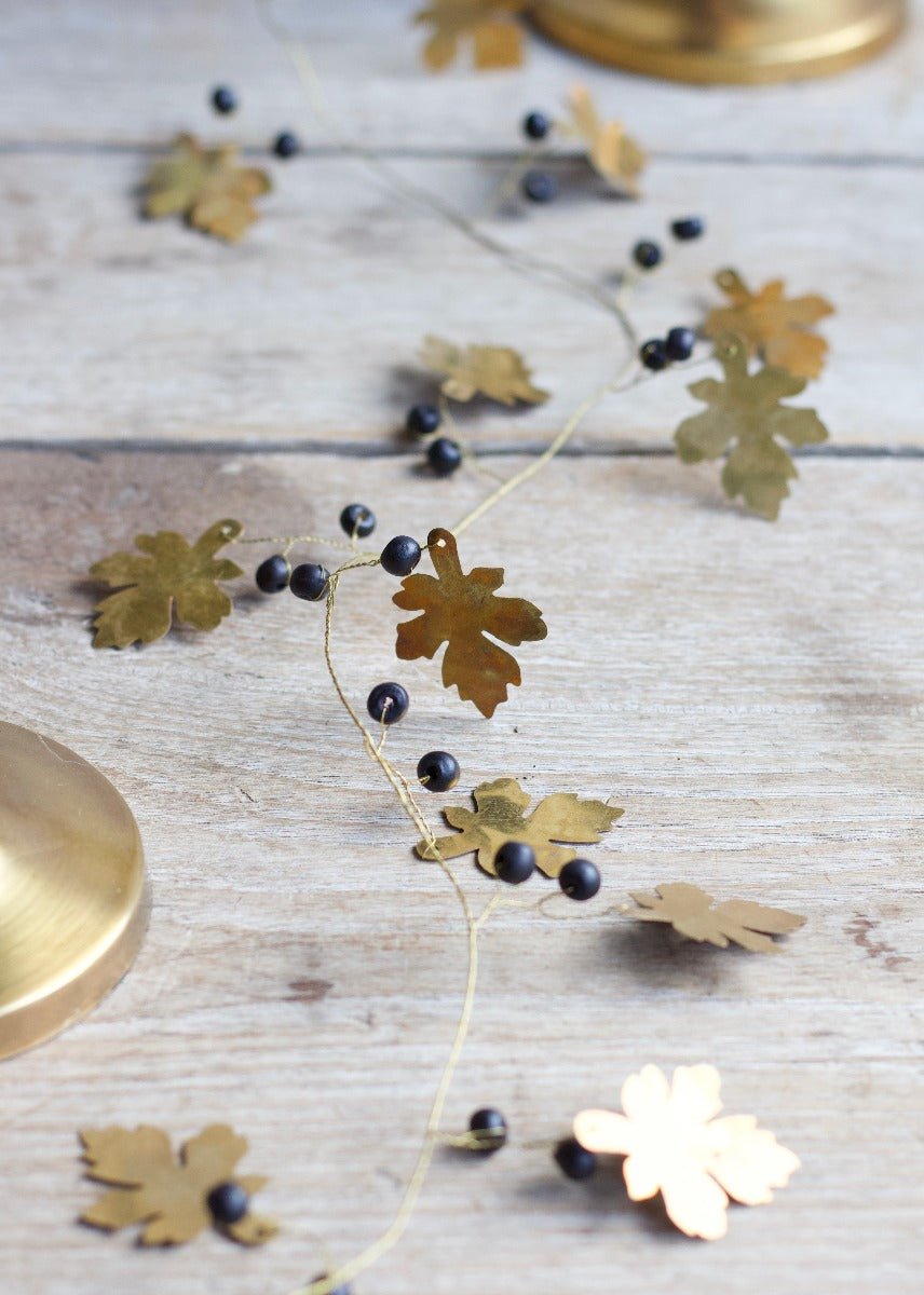Close-up of a decorative garland with golden brass oak leaves and small black beads, arranged on a rustic wooden surface with a brass object partially visible in the corner.