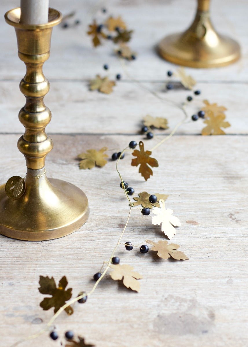Brass garland with oak-shaped golden leaves and small black beads, styled across a wooden surface between two golden candleholders with white taper candles.