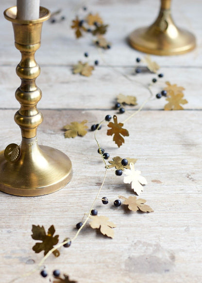 Brass garland with oak-shaped golden leaves and small black beads, styled across a wooden surface between two golden candleholders with white taper candles.
