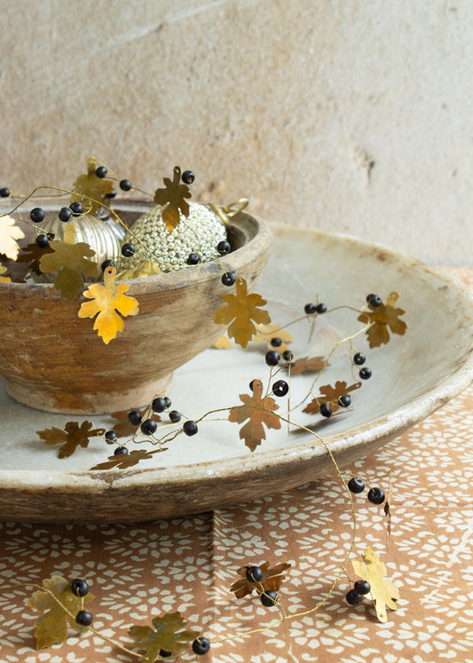 Garland with brass oak leaves and small black berries draped across a rustic bowl filled with ornaments, placed on a stone plate with a patterned cloth underneath.