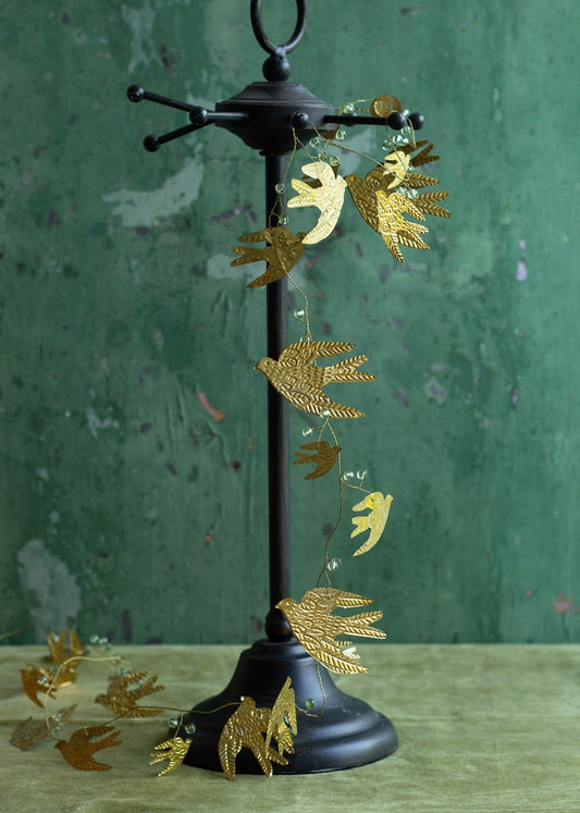 Garland with embossed golden birds and green glass beads, wrapped around a black metal stand against a textured green wall with a velvet-like green surface.