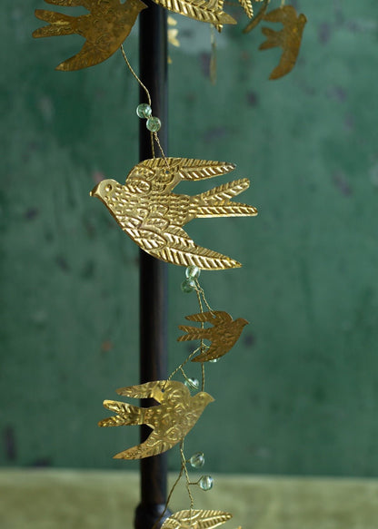 Close-up of a decorative garland with detailed golden brass birds and translucent green glass beads, hanging against a deep green textured wall.