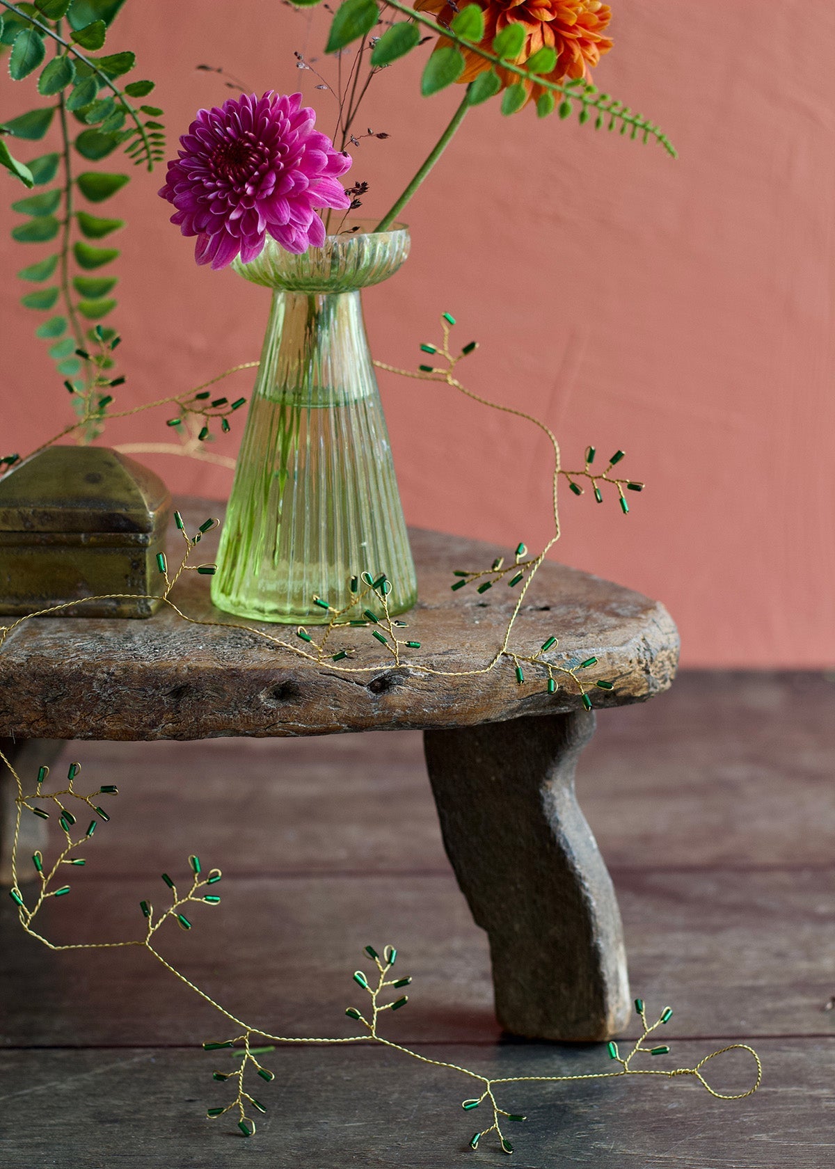 Golden wire garland with small jade-green glass beads draped across a rustic wooden table beside a glass vase with flowers.