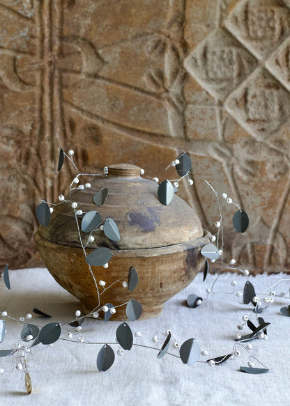 Garland with matte grey metal leaves and white beads, styled around a rustic wooden pot with a textured background.