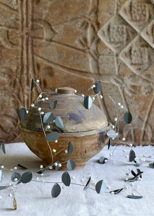 Garland with matte grey metal leaves and white beads, styled around a rustic wooden pot with a textured background.