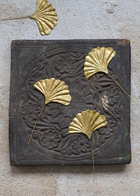 Golden brass ginkgo leaf ornaments with embossed details, displayed on a dark wooden plate.