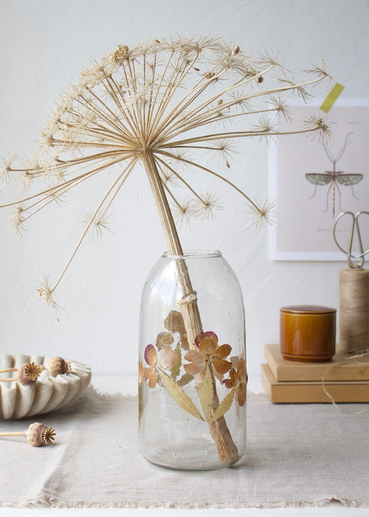 Clear glass vase with real pressed flowers and a dried allium stem on a linen-covered table.