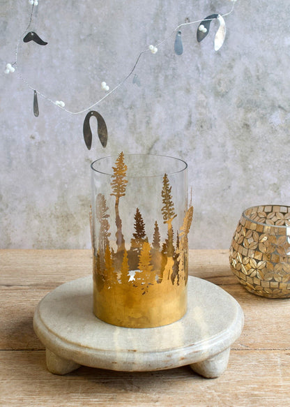 Glass candle holder with gold forest tree cutouts placed on a stone stand.