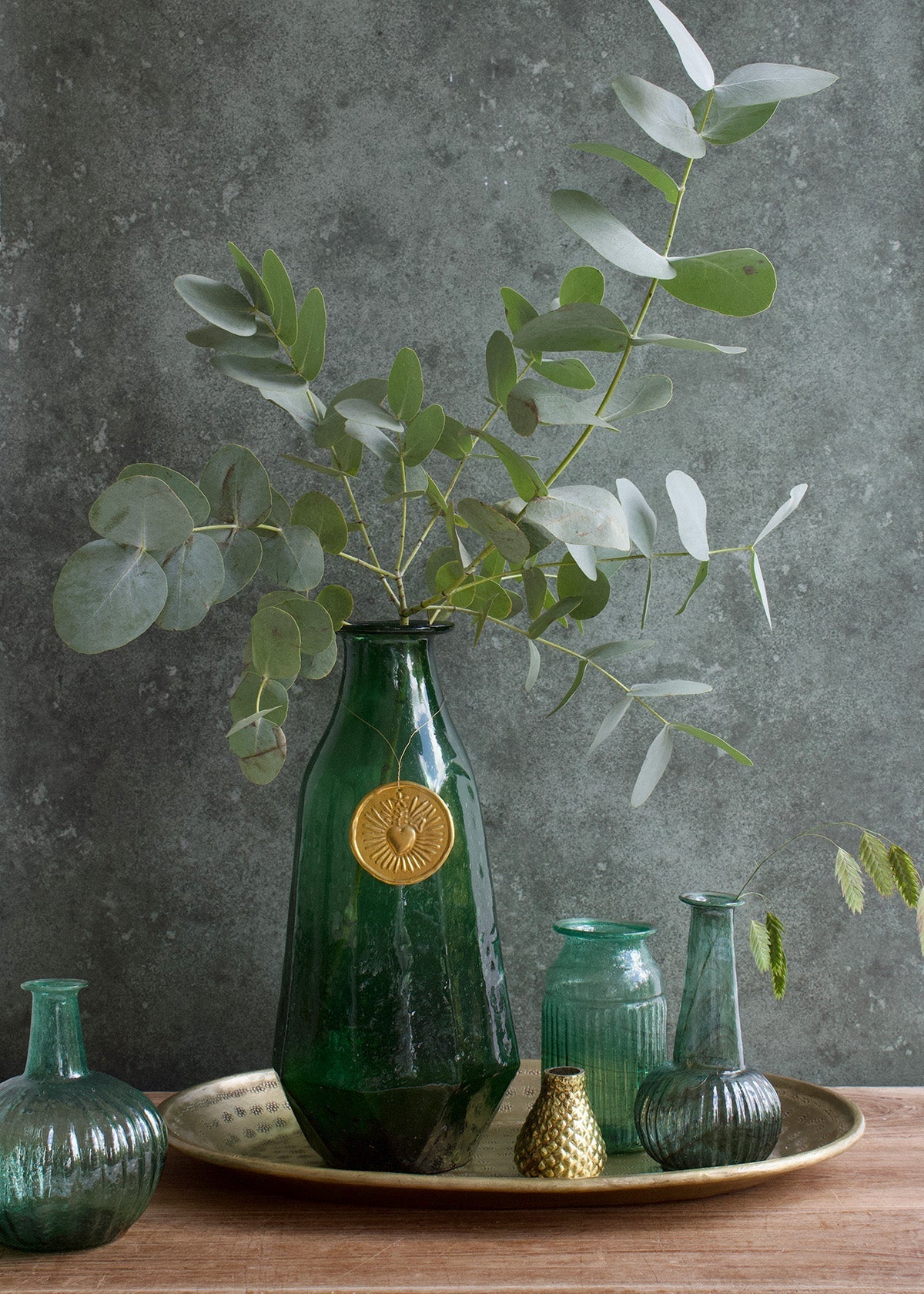 Green vase with gold emblem on a tray with eucalyptus leaves against a gray background