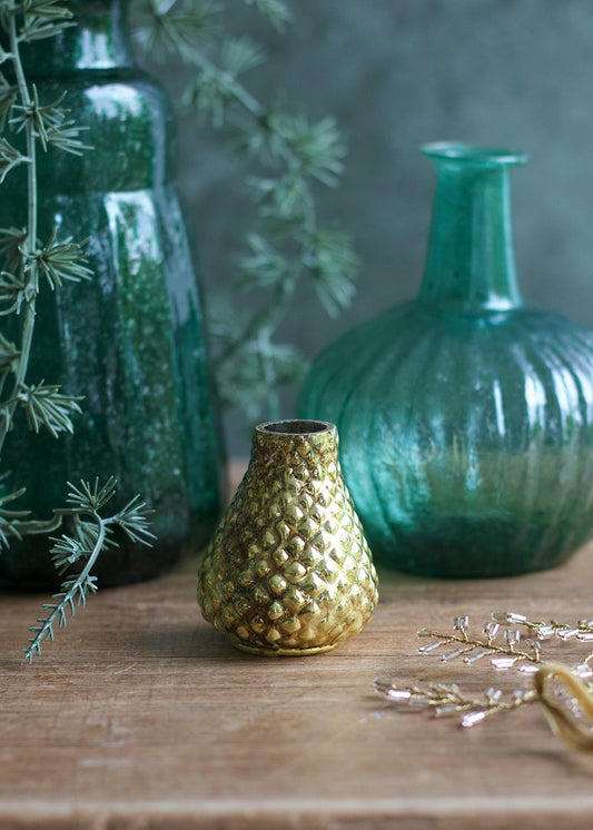 Gold textured vase on a wooden surface with green textured vases and plants in the background.