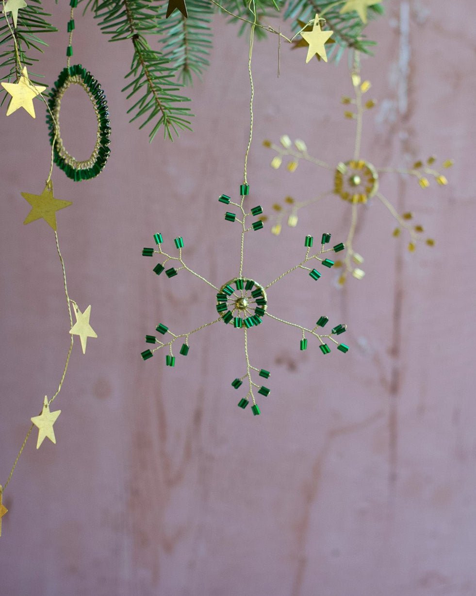 Handmade green and gold glass bead snowflake ornament hanging on a fir branch.