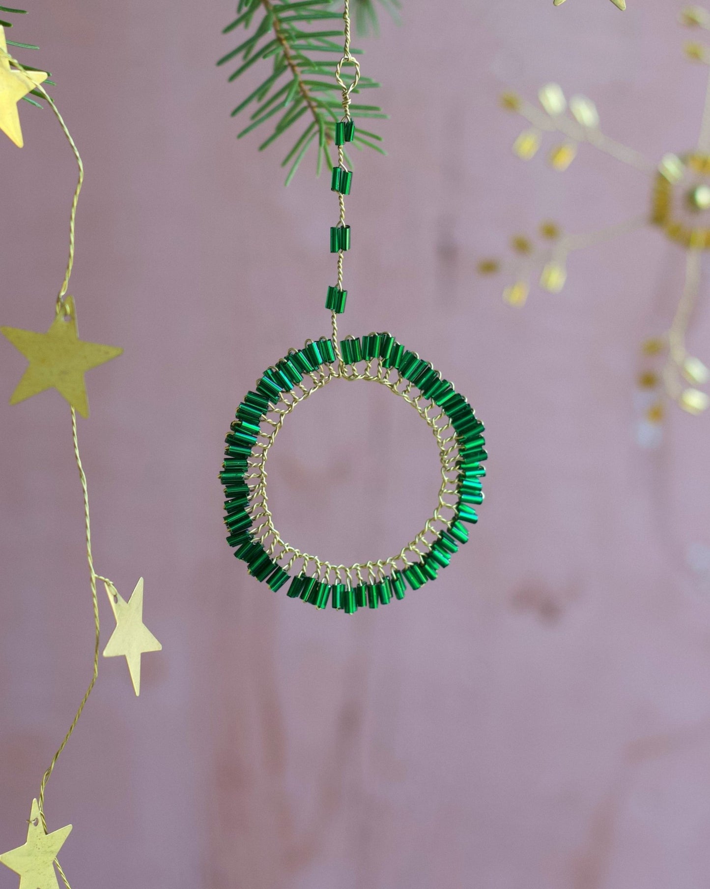 Decorative green beaded circle and yellow star ornaments hanging from a branch against a pink background.