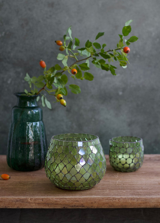 Green glass vases with a textured pattern on a wooden surface, featuring a branch with leaves and small fruits against a dark background.