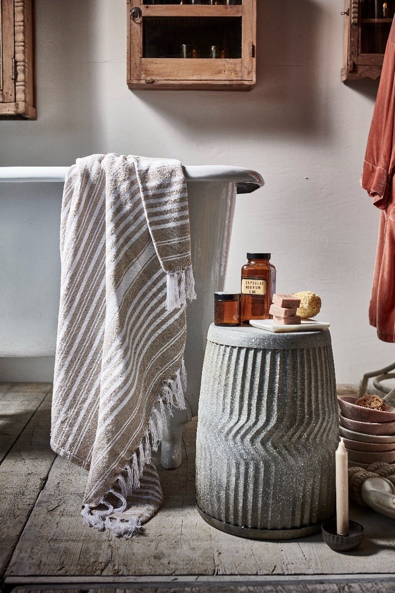 Bathroom setting with a striped towel draped over a bathtub, a textured stone stool with bottles and candles, and wooden cabinets.