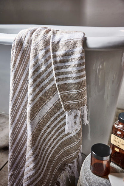 Striped towel hanging over a bathtub with a bottle and glass on a ledge.