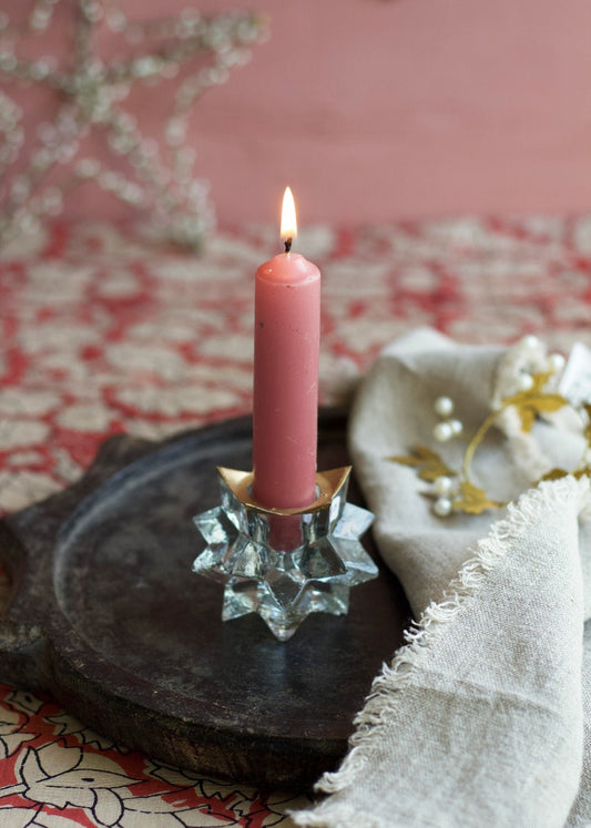 Clear glass star-shaped candle holder with painted gold accent, holding a lit pink taper candle on a dark wooden tray with soft fabric nearby.