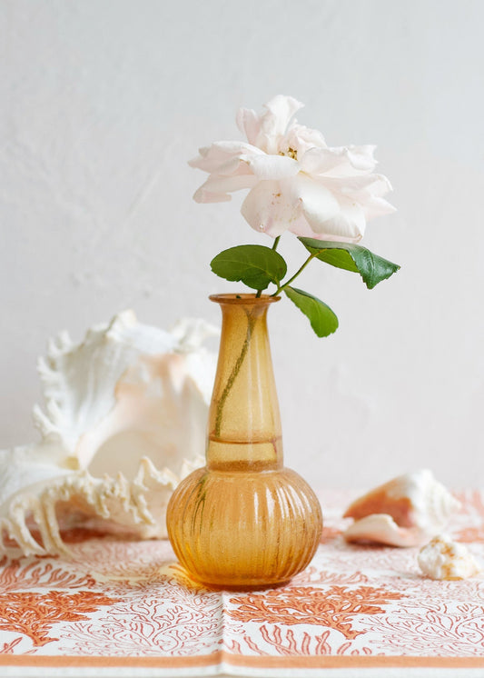 Small amber glass bud vase with ribbed base and long neck, holding a single pale rose on a patterned fabric with seashells.