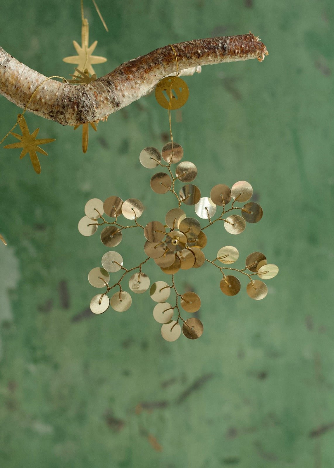 Handmade brass-colored snowflake ornament made of flat paillettes, hanging from a branch and styled on a napkin for a festive table setting.