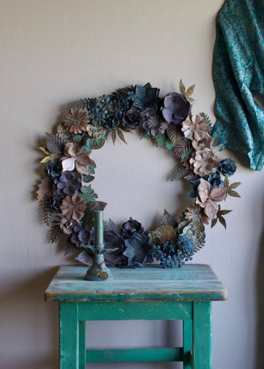 Large decorative wreath made of handcrafted metal flowers in blue, green, and neutral tones, hanging on a wall.
