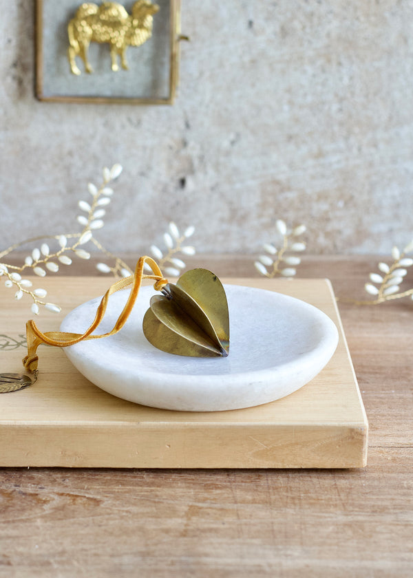 Three-dimensional brass heart ornament with velvet ribbon, displayed on a white marble dish.