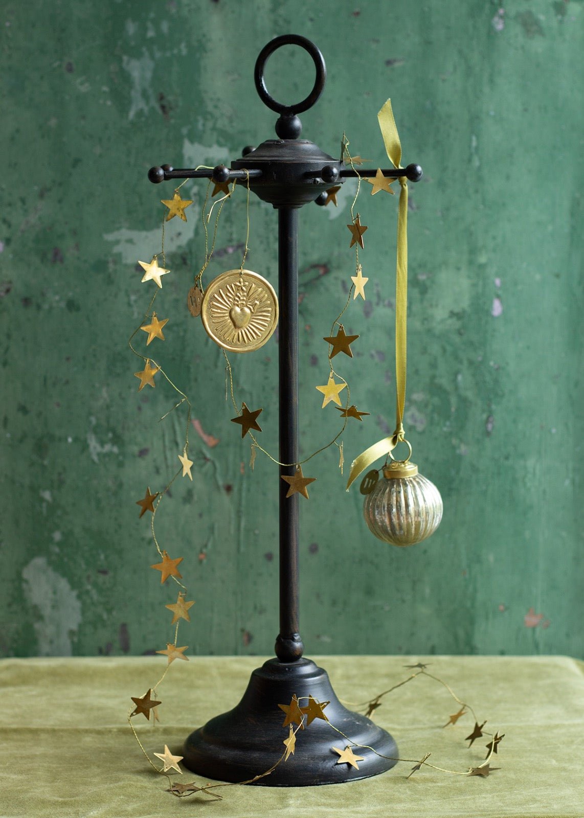 Holiday ornament display with a brass Sacred Heart medallion and vintage glass bauble hanging from a black metal stand, decorated with a golden star garland, set against a textured green background.