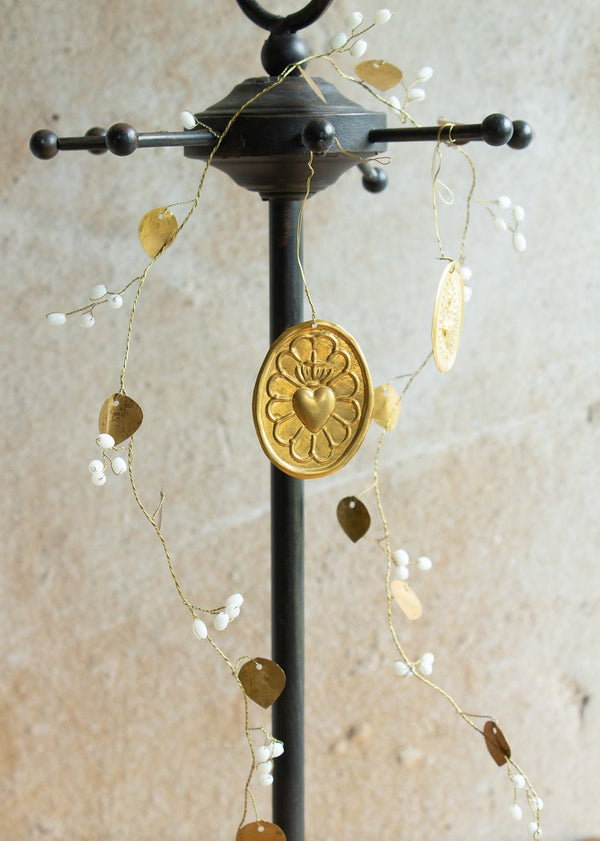 Gold-colored metal heart pendant with crown and petal-like frame, hanging from a garland with white beads and brass leaves.