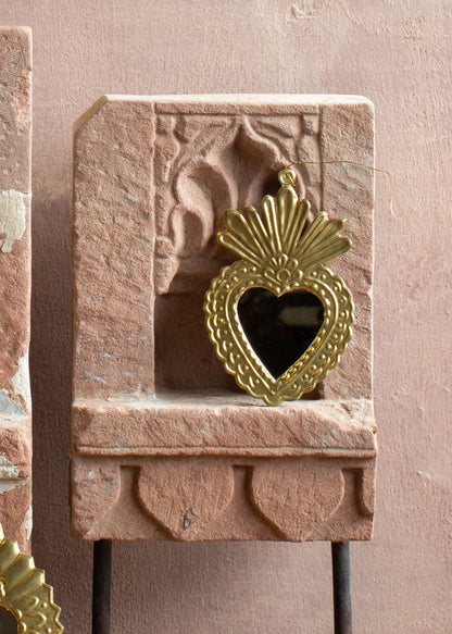 Brass ex-voto ornament with a small heart-shaped mirror and embossed details, displayed against stone.