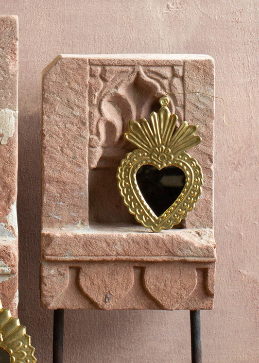 Brass ex-voto ornament with a small heart-shaped mirror and embossed details, displayed against stone.