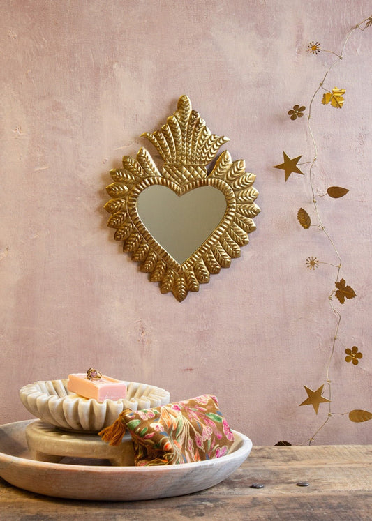Golden heart-shaped ex-voto mirror with radiant embossed detailing, hanging on a soft pink textured wall, styled with a wooden table and floral accessories.