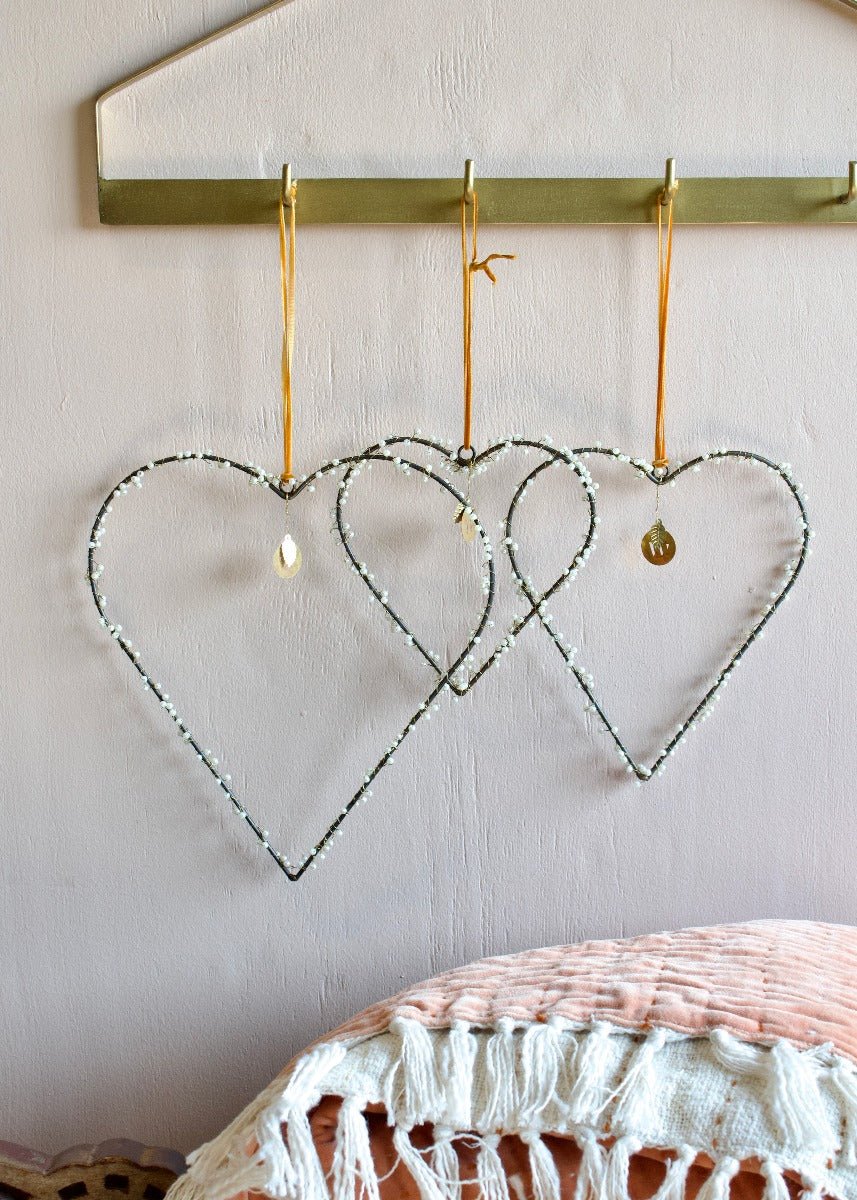 Three pearl-beaded heart ornaments hanging from gold hooks against a soft pink wall